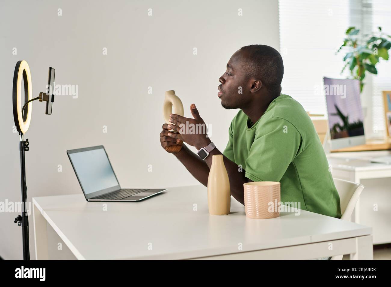 Junger afroamerikanischer Mann mit handgefertigter Vase in der Hand, der mit dem Online-Publikum spricht, während er während des Livestreams in der Smartphone-Kamera schaut Stockfoto