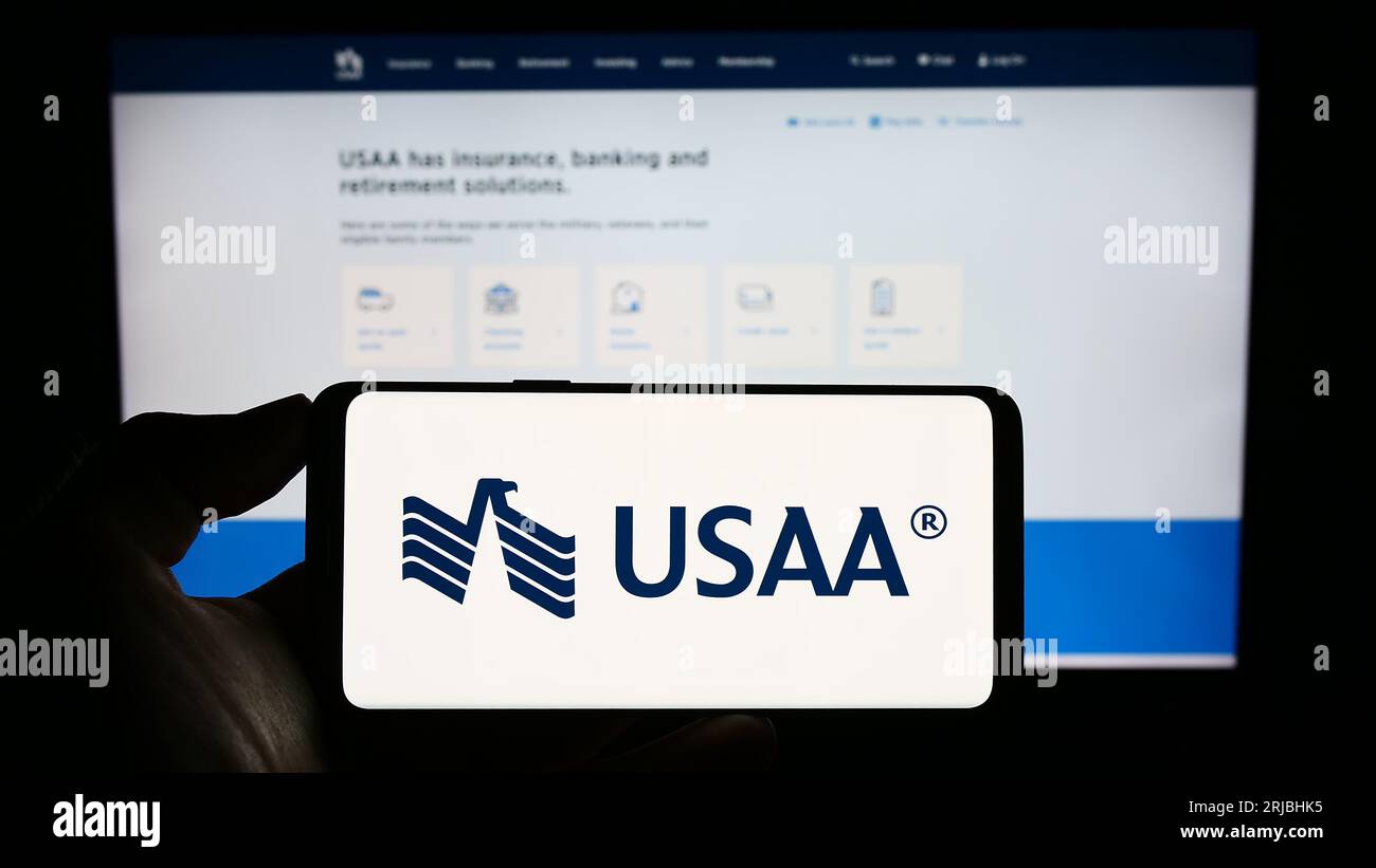 Person holding cellphone with logo of United Services Automobile Association (USAA) on screen in front of webpage. Focus on phone display. Stockfoto