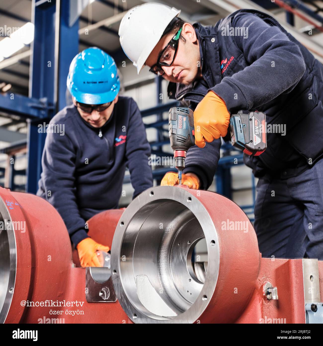 Zwei Männer, die an einem Metallrohr arbeiten und einen Bohrer in einer Fabrik benutzen, während sie Arbeitskleidung, Schutzbrille und Helme tragen. Stockfoto
