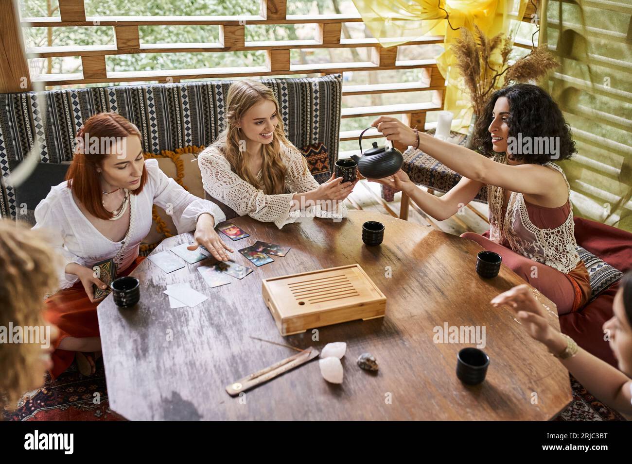 Multirassische Frau, die Tee in der Nähe von Freundinnen gießt, die Tarotkarten im Retreat-Center lesen Stockfoto