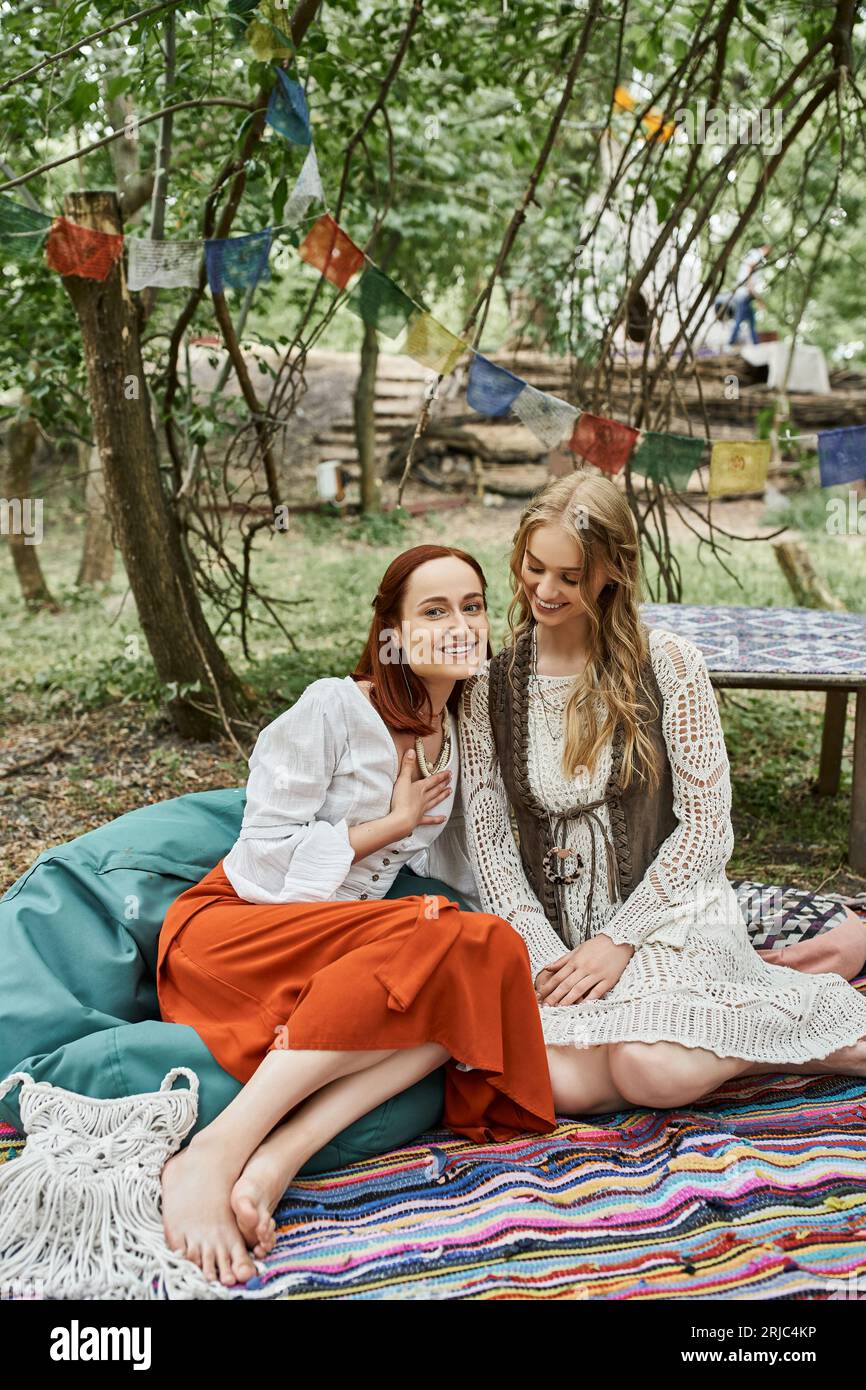 Positive rothaarige Frau in Boho-Kleidung sitzt in der Nähe eines Freundes auf einer Wiese im Retreat Center Stockfoto