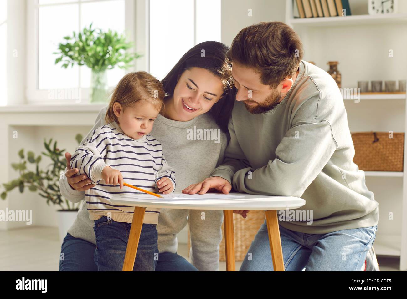 Das junge Ehepaar sitzt zusammen mit seiner Tochter auf dem Boden und zeichnet mit einem Bleistift auf ein weißes Blatt Papier Stockfoto