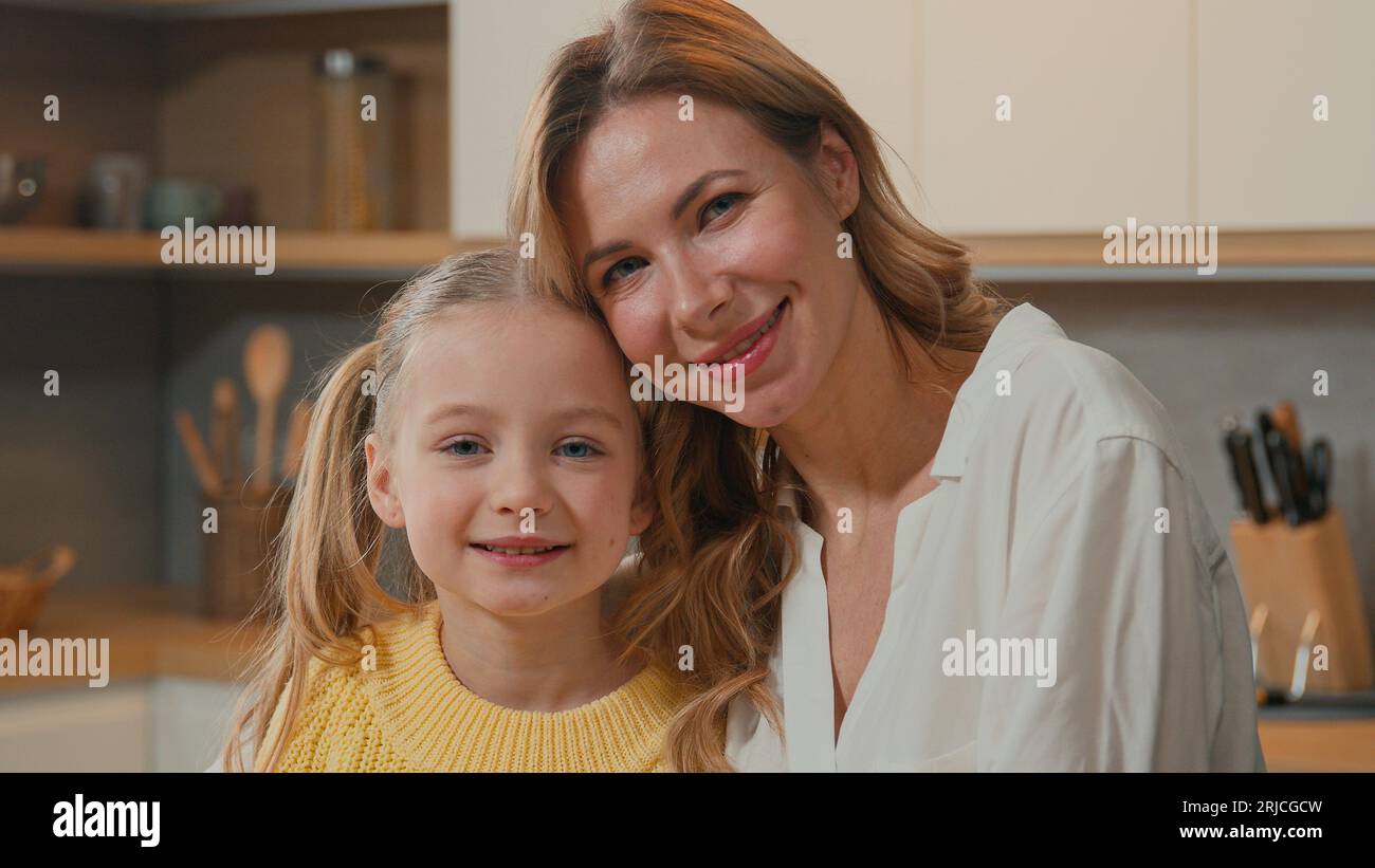 Kaukasische glückliche Familie in der Küche. Nahaufnahme lächelndes Kind Tochter kuschelnde Mutter Blick in die Kamera. Erwachsene alleinerziehende Mutter pflegt Eltern und adoptiert süßes Kind Stockfoto