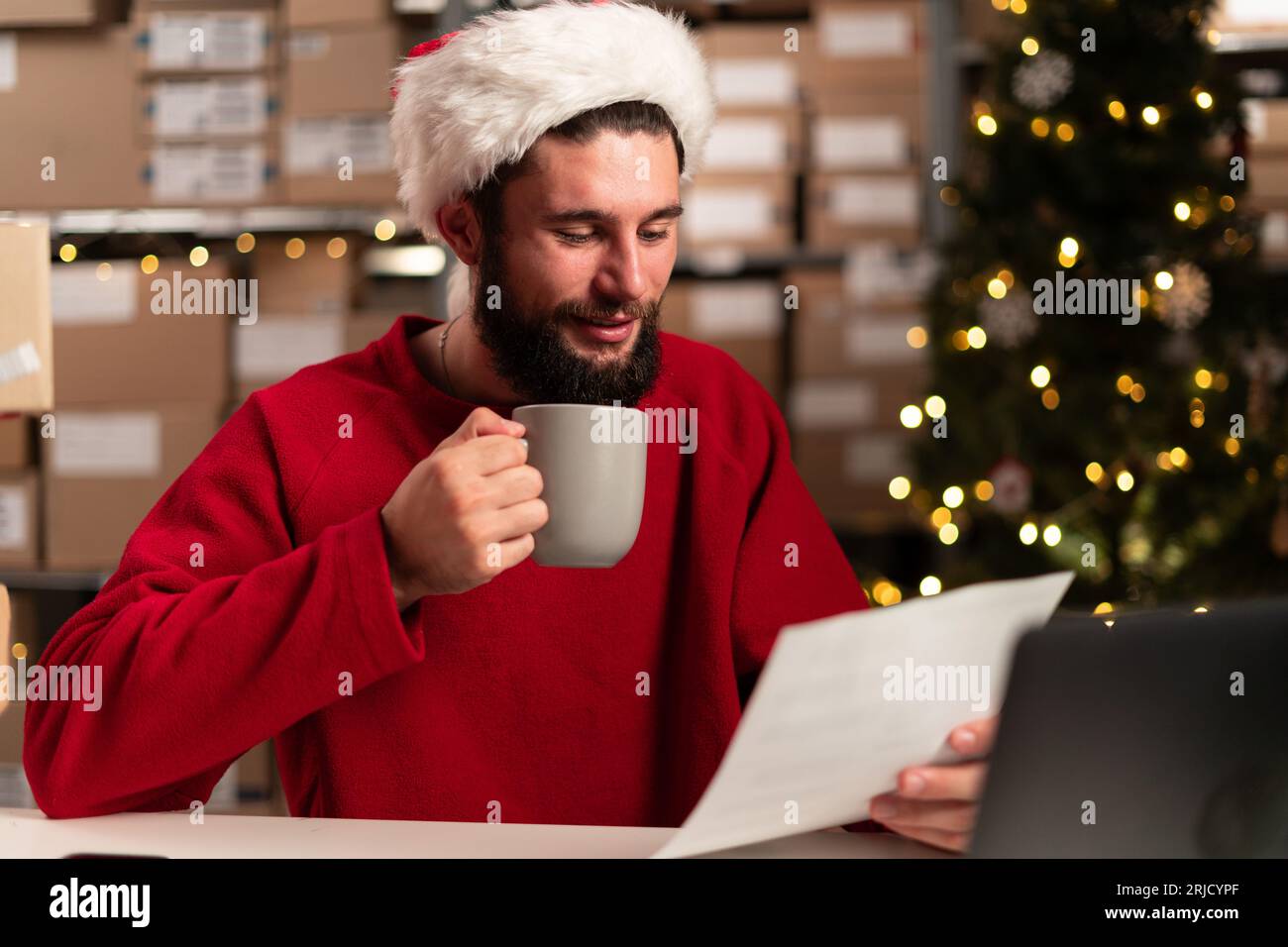 Weihnachtsgeschäft. Happy Modern Wear Santa Hut mit Laptop und Paketen mit Dokumenten und Kaffee im Lager. Stockfoto