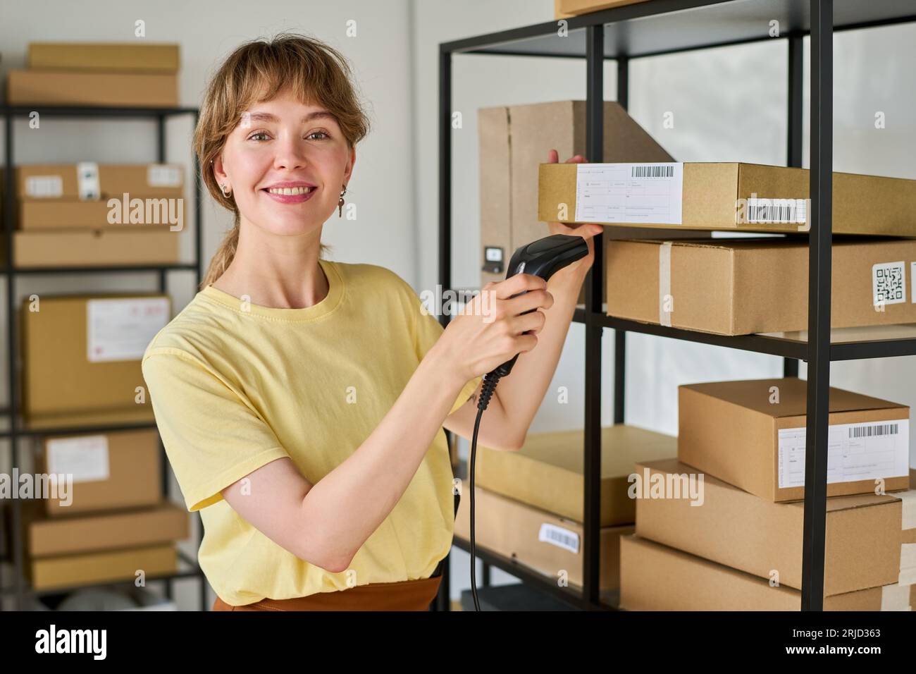 Glückliche junge Managerin von Marketplace mit Scanner, der die Kamera anschaut, während er qr-Codes auf verpackten Kartons mit bestellten Waren überprüft Stockfoto