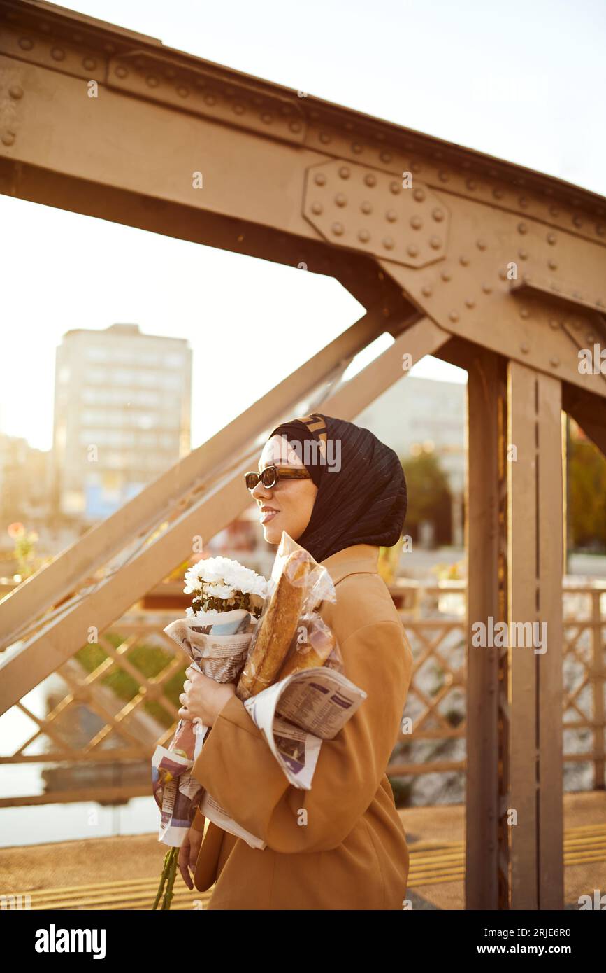 Eine Hidschab-Frau in stilvoller Sonnenbrille und einem eleganten französischen Outfit, die bei Sonnenuntergang durch die Stadt spaziert, mit einem Blumenstrauß, Brot und einer Zeitung Stockfoto