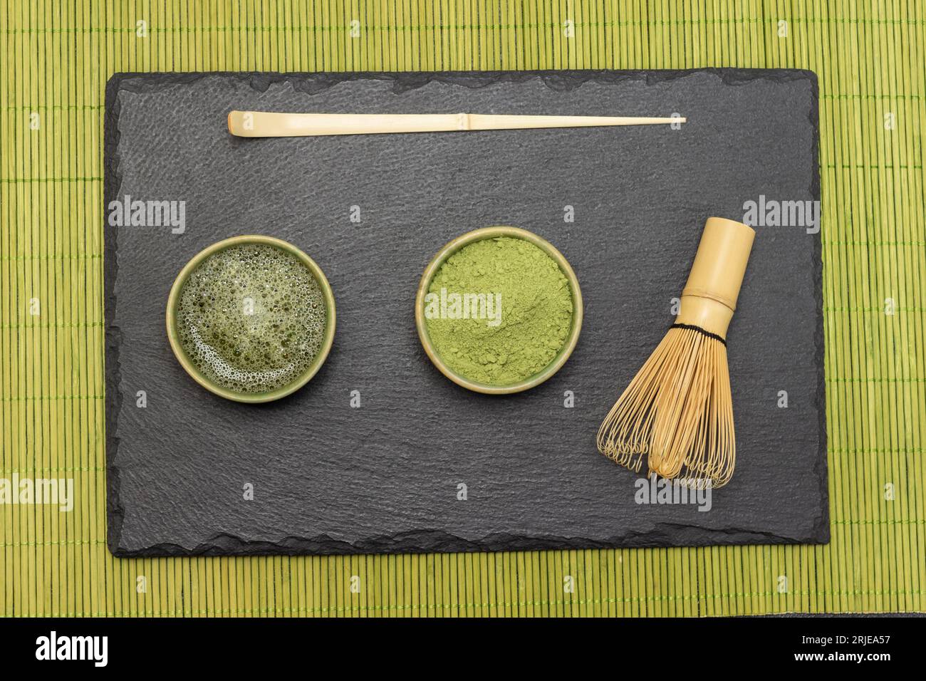 Teelöffel, Bambusbesen, Matcha-Teepulver und Matcha-Tee auf Steinbrett. Flach liegend. Hintergrund von kleinen grünen Holzstäben Stockfoto