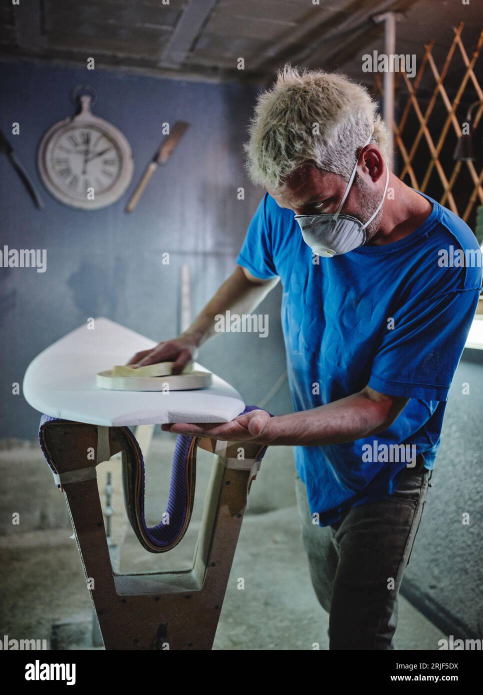 Seitenansicht des konzentrierten blonden männlichen Meisters in der Atemschutzmaske, der weißes Surfbrett poliert, das in der Werkstatt auf Gestellen platziert wird Stockfoto