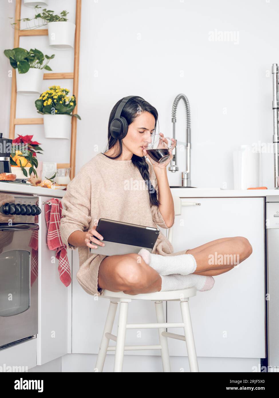 Ganzer Körper einer jungen hispanischen Frau in Kopfhörern, die aromatischen frischen Kaffee trinkt und sich interessante Filme ansieht, während sie mit überkreuzten Beinen sitzt Stockfoto