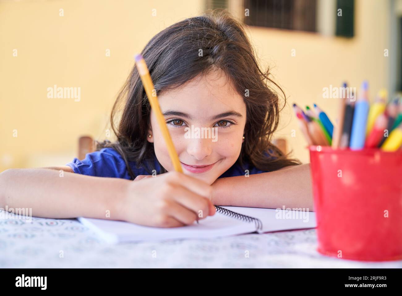 Kreatives, süßes Schulmädchen, das Notizen auf Papier mit Bleistift im Copybook macht, während es Zeit zu Hause verbringt und die Kamera betrachtet Stockfoto