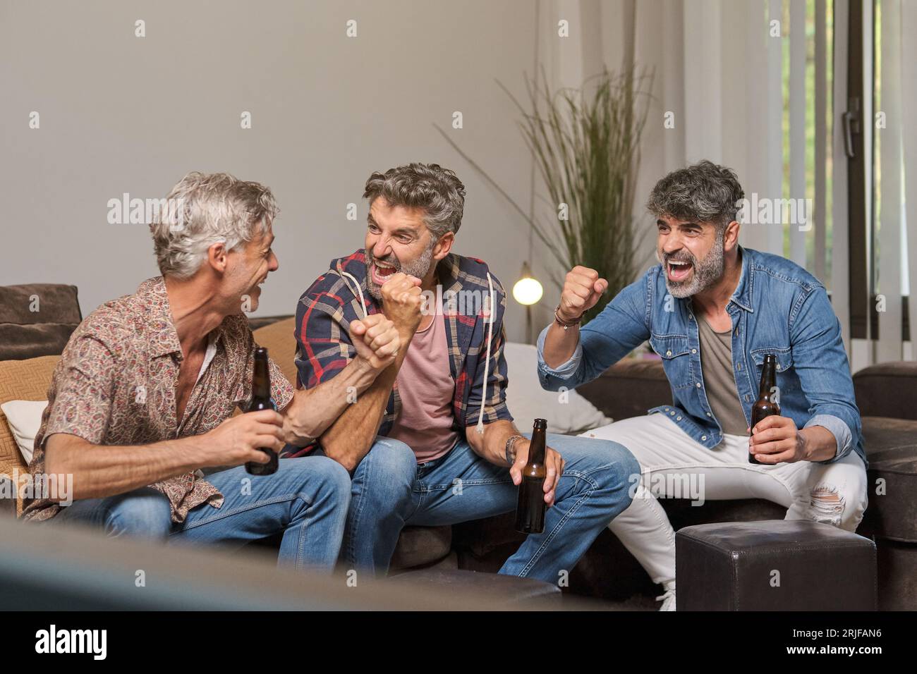Erwachsene Männer schauen sich an und feiern das Tor im Spiel, während sie auf dem Sofa sitzen und zu Hause Bier trinken Stockfoto