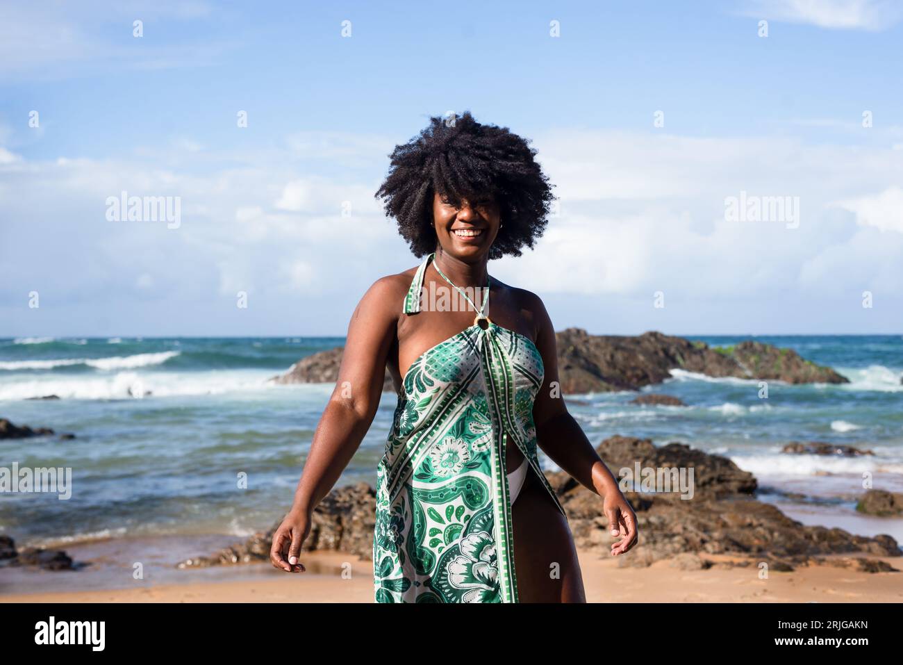 Porträt einer wunderschönen, schwarzhaarigen Frau, die lächelt und glücklich am Strand steht. Im Hintergrund liegen die Wolken auf Felsen und das Meer. Rio Vermelho, Salvad Stockfoto