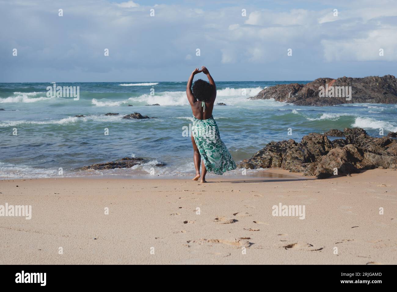 Das lange Porträt einer wunderschönen Frau mit schwarzem, kraftvollem Haar, die mit dem Rücken am Strand stand, bewegte sich mit ihren Armen. Die Wolken rocken Stockfoto