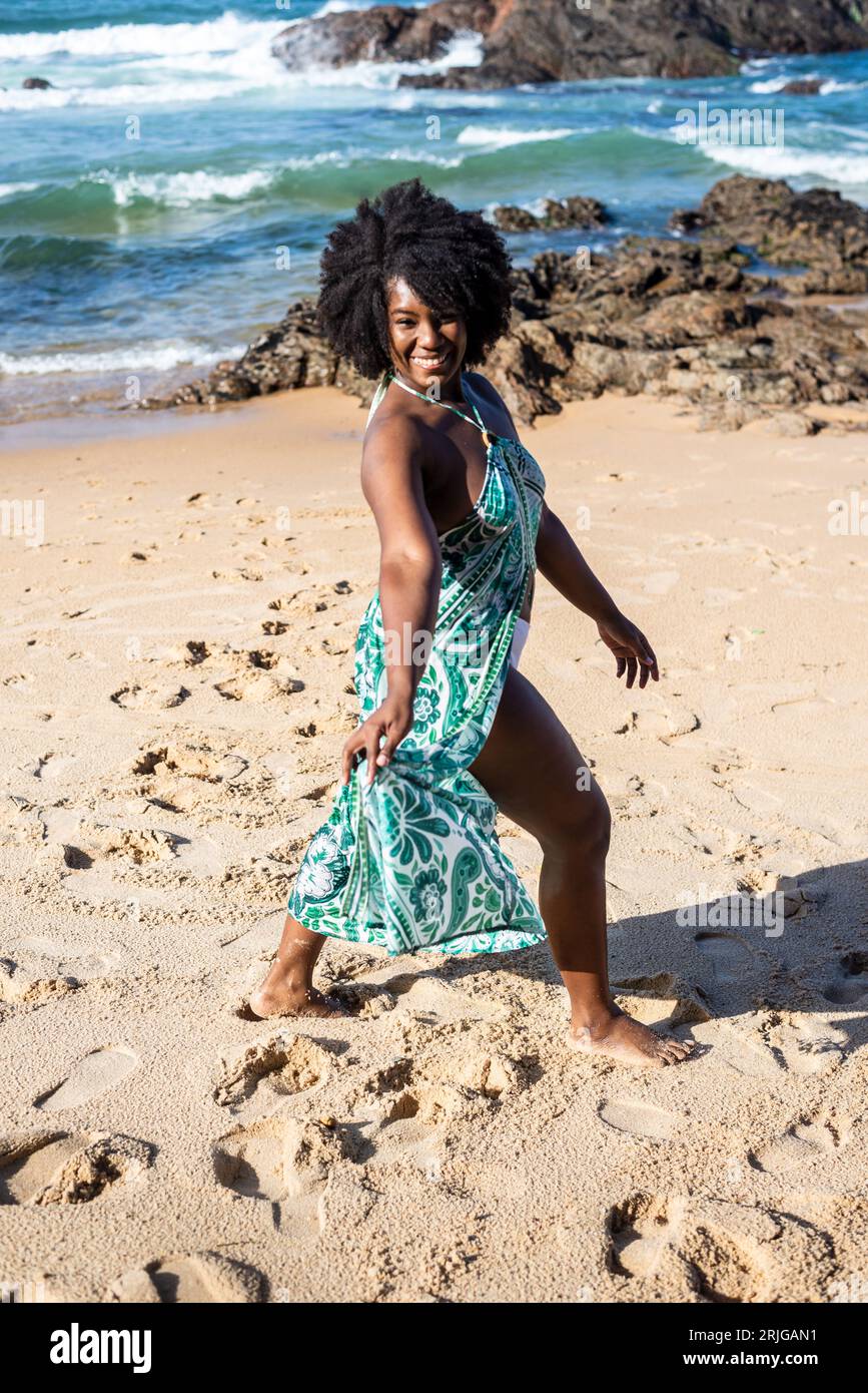 Eine wunderschöne Frau mit schwarzem, kräftigem Haar, die ein blaues Kleid trägt und im Sand des Strandes spielt. Glückliche und selbstbewusste Person. Im Hintergrund der Himmel Stockfoto