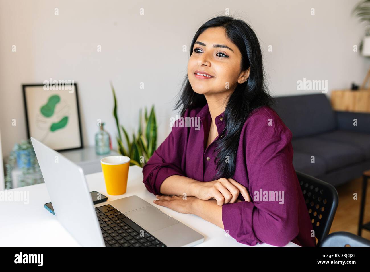 Nachdenkliche indische Frau, die mit Laptop auf dem Tisch sitzt, während sie wegschaut Stockfoto