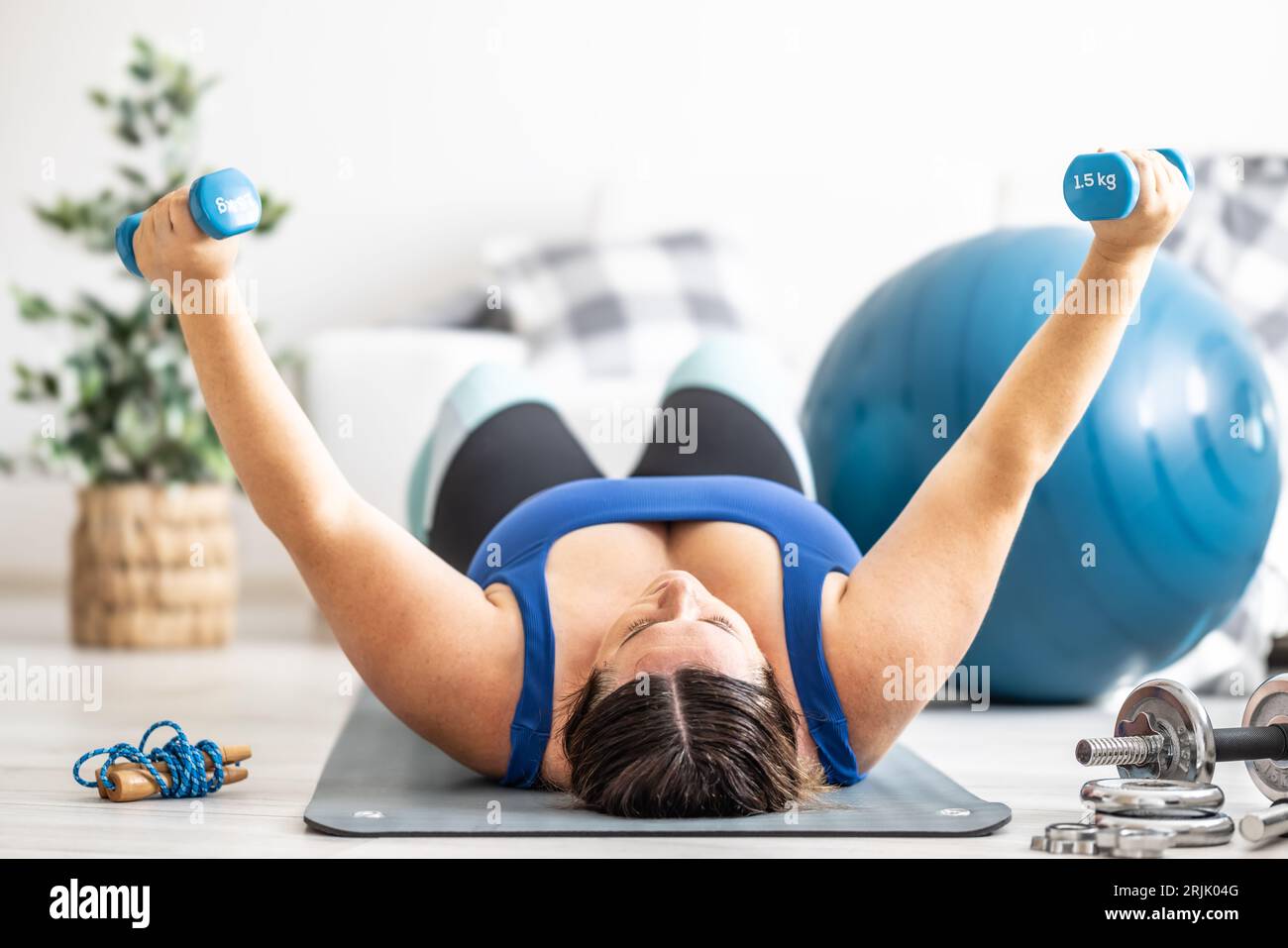 Übergewichtige Dame, die im Wohnzimmer Sport treibt. Er liegt auf der Matte und hebt die Kurzhanteln. Stockfoto