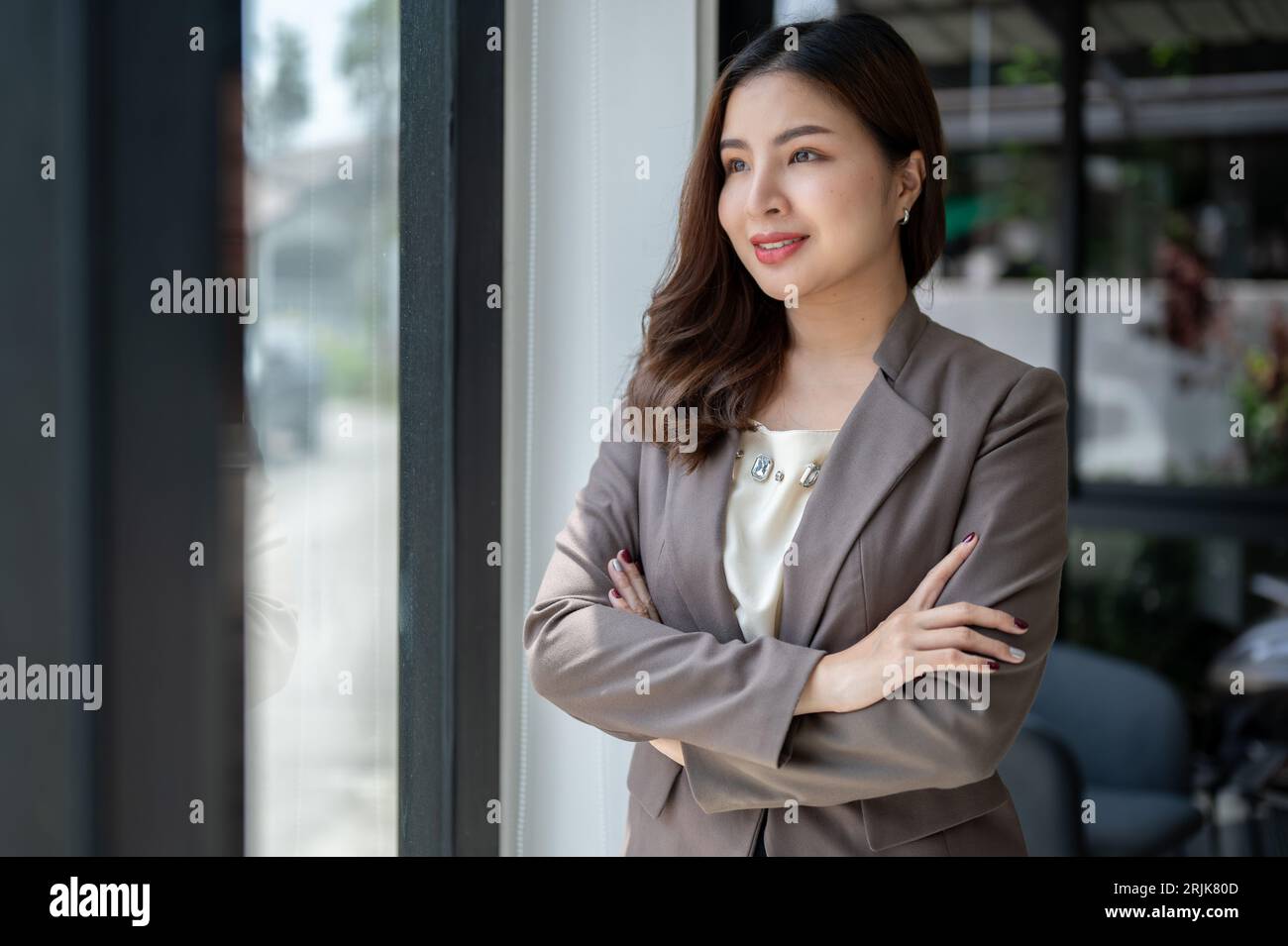 Eine wunderschöne, wunderschöne, tausendjährige asiatische Geschäftsfrau oder CEO in einem formellen Business-Anzug steht mit überkreuzten Armen, träumt und schaut aus Stockfoto