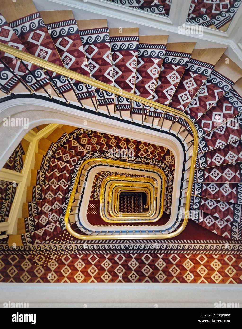 Luxuriöse Treppe in abstrakter Form mit roter Teppichabdeckung und goldenem Handlauf. Hypnotische Muster-Wendeltreppe Stockfoto