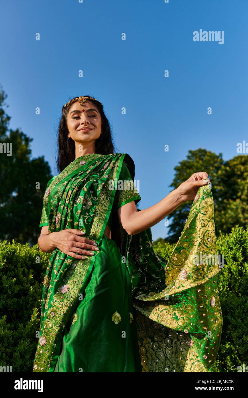 Fröhliche und moderne junge indische Frau berührt Sari mit Muster in der Nähe von grünen Pflanzen im Park draußen Stockfoto