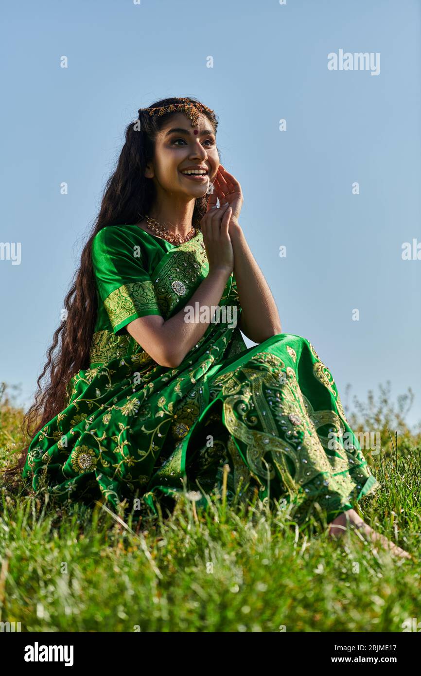 Fröhliche indianerin in Sari und matha patti, die auf einem grasbewachsenen Hügel mit blauem Himmel im Hintergrund sitzen Stockfoto