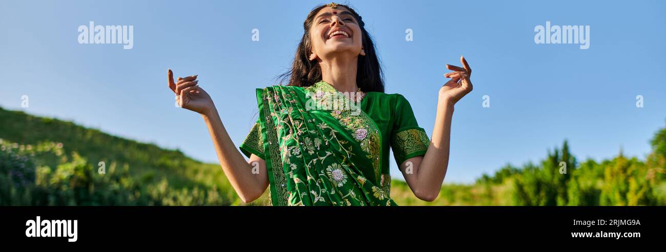 Glückliche junge indische Frau in grünem Sari stehend mit Sommerlandschaft auf Hintergrund, Banner Stockfoto