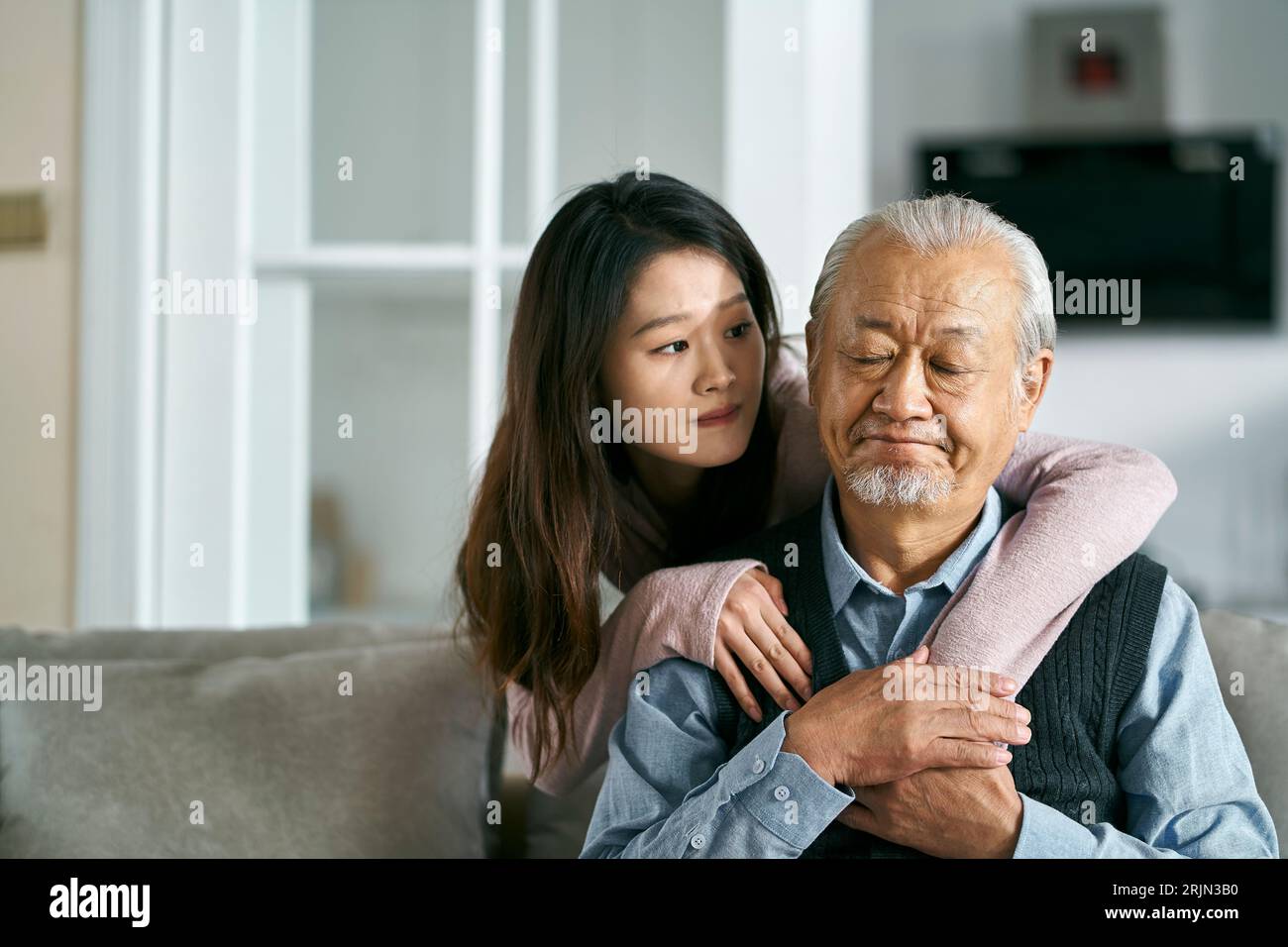 Trauriger älterer asiatischer Vater, der auf der Couch im Wohnzimmer zu Hause saß, getröstet von einer erwachsenen Tochter Stockfoto