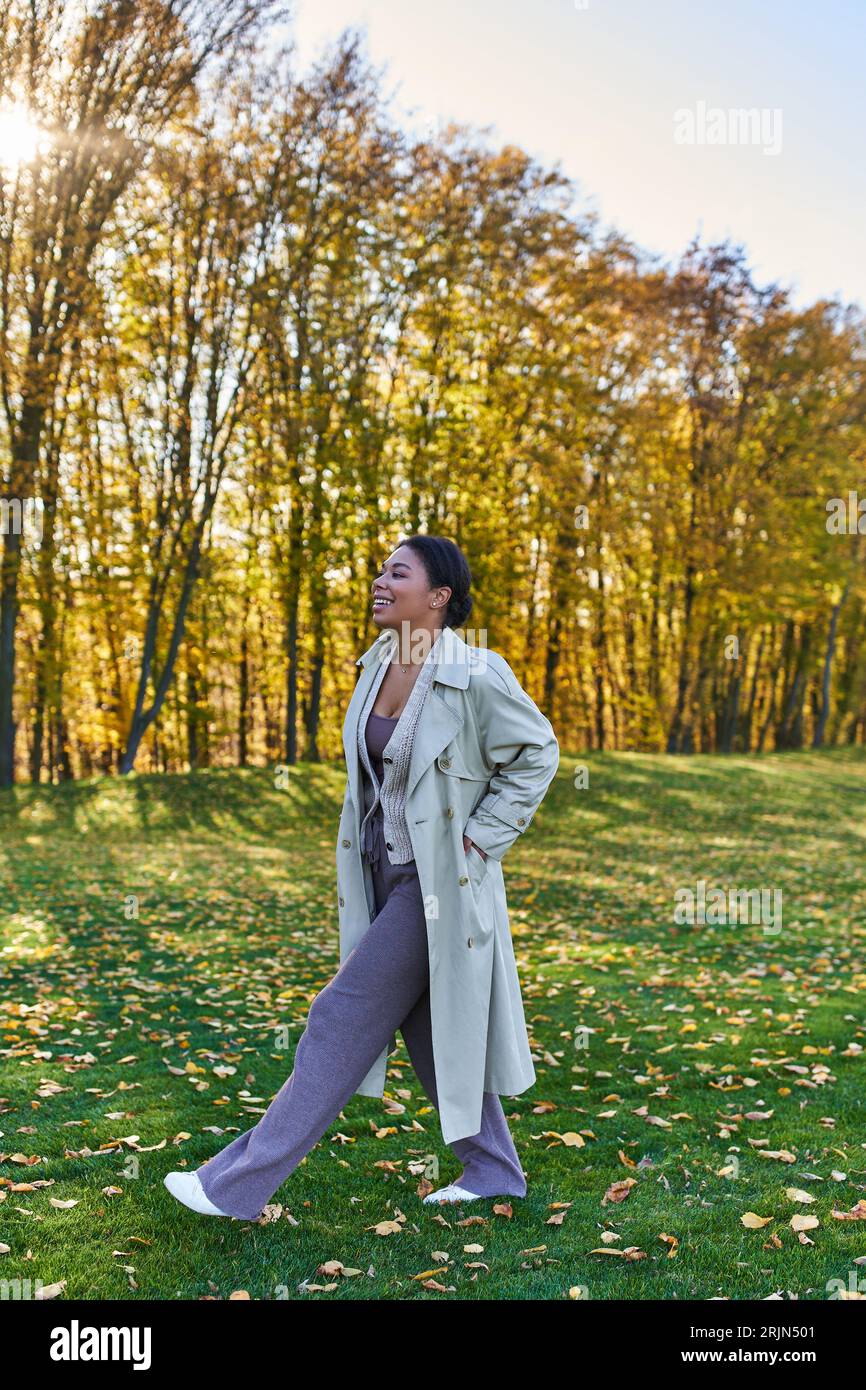 Glückliche afroamerikanische Frau im Grabenmantel, die auf Gras mit gefallenen Blättern geht, Herbst, Mode Stockfoto