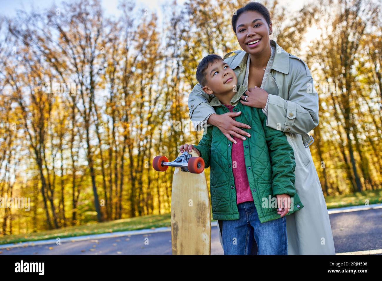 Glückliche afroamerikanische Frau umarmt Sohn mit Penny Board, Herbst, Herbstsaison, Mutterliebe, Park Stockfoto