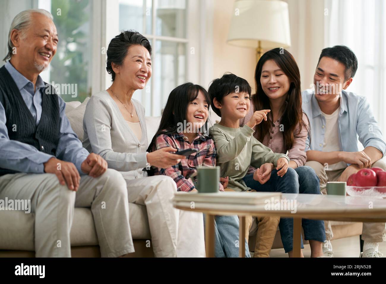 Mehrgenerationenübergreifende asiatische Familie, die im Wohnzimmer auf der Couch sitzt und sich die Fernsehserie glücklich und lächelnd ansieht Stockfoto