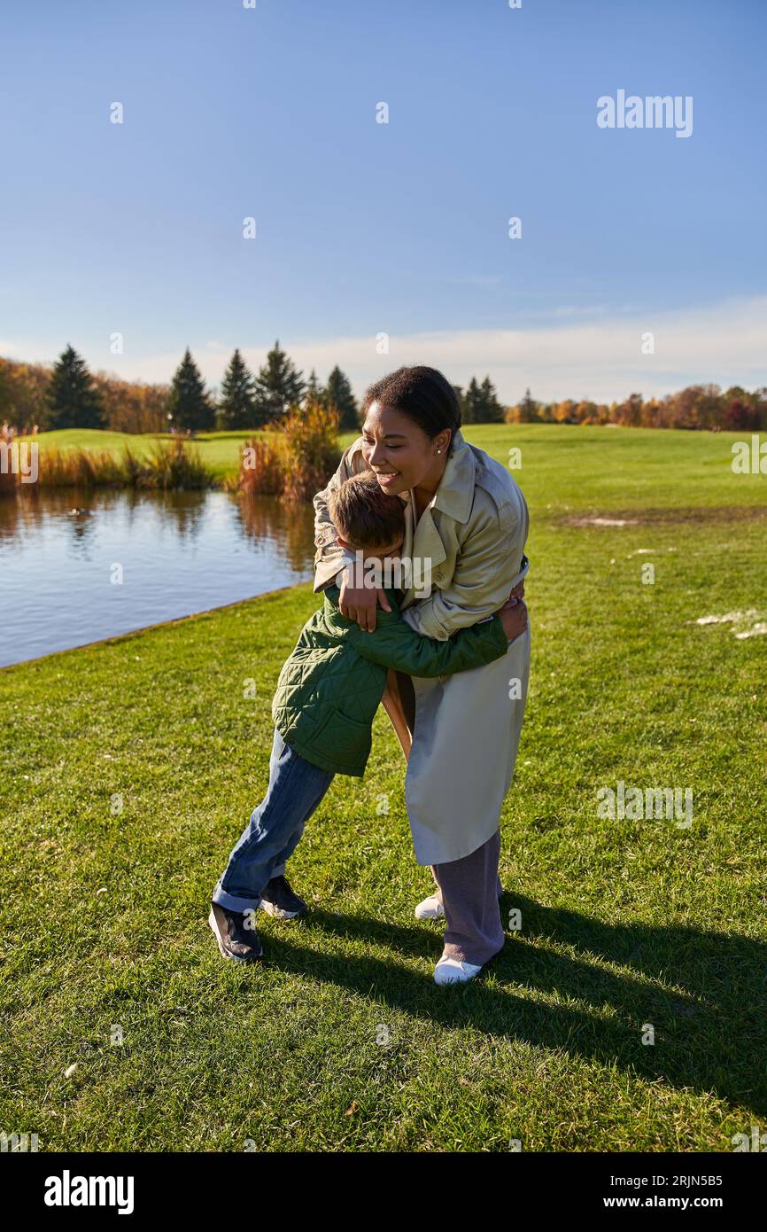 Glückliche Frau umarmt Sohn, steht in der Nähe des Sees im Park, afroamerikanische Familie, Herbstmode Stockfoto