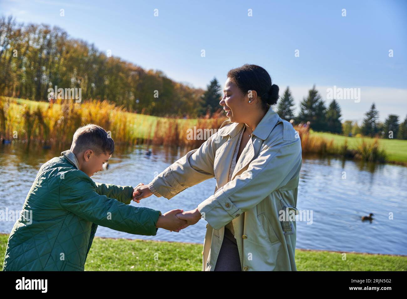 Glückliche afroamerikanische Mutter hält Hände mit Sohn in der Nähe von Teich, Herbstsaison, Familienbindung, Lachen Stockfoto