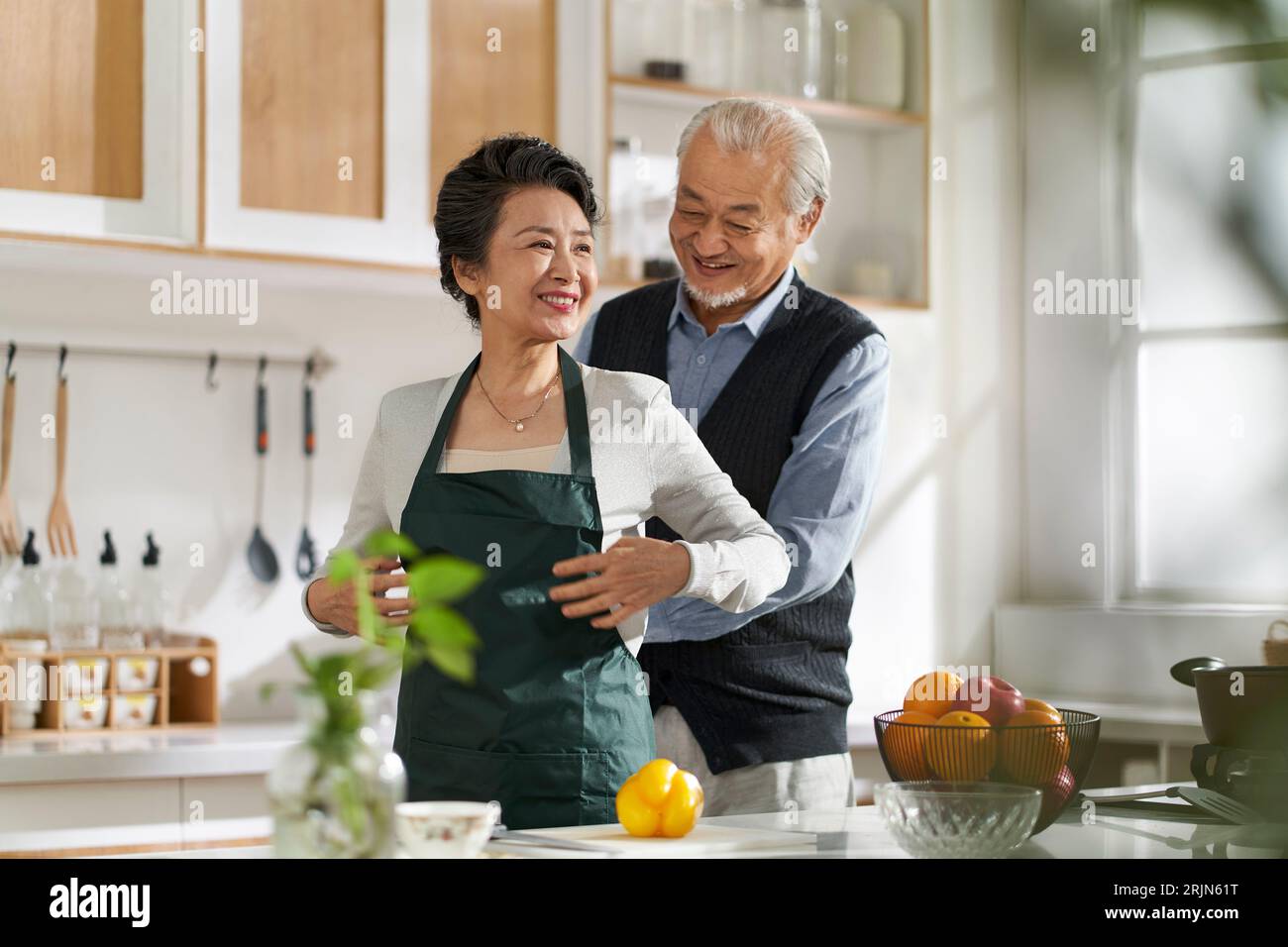 Liebender älterer asiatischer Mann Ehemann, der Frau hilft, die Schürze in der Küche zu Hause zu binden Stockfoto