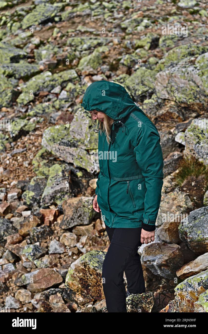 Porträt einer lächelnden Wanderin in grüner Jacke und schwarzer Reisehose, die auf einem felsigen Berghang spaziert. Reise- und aktives Lifestyle-Konzept. Stockfoto