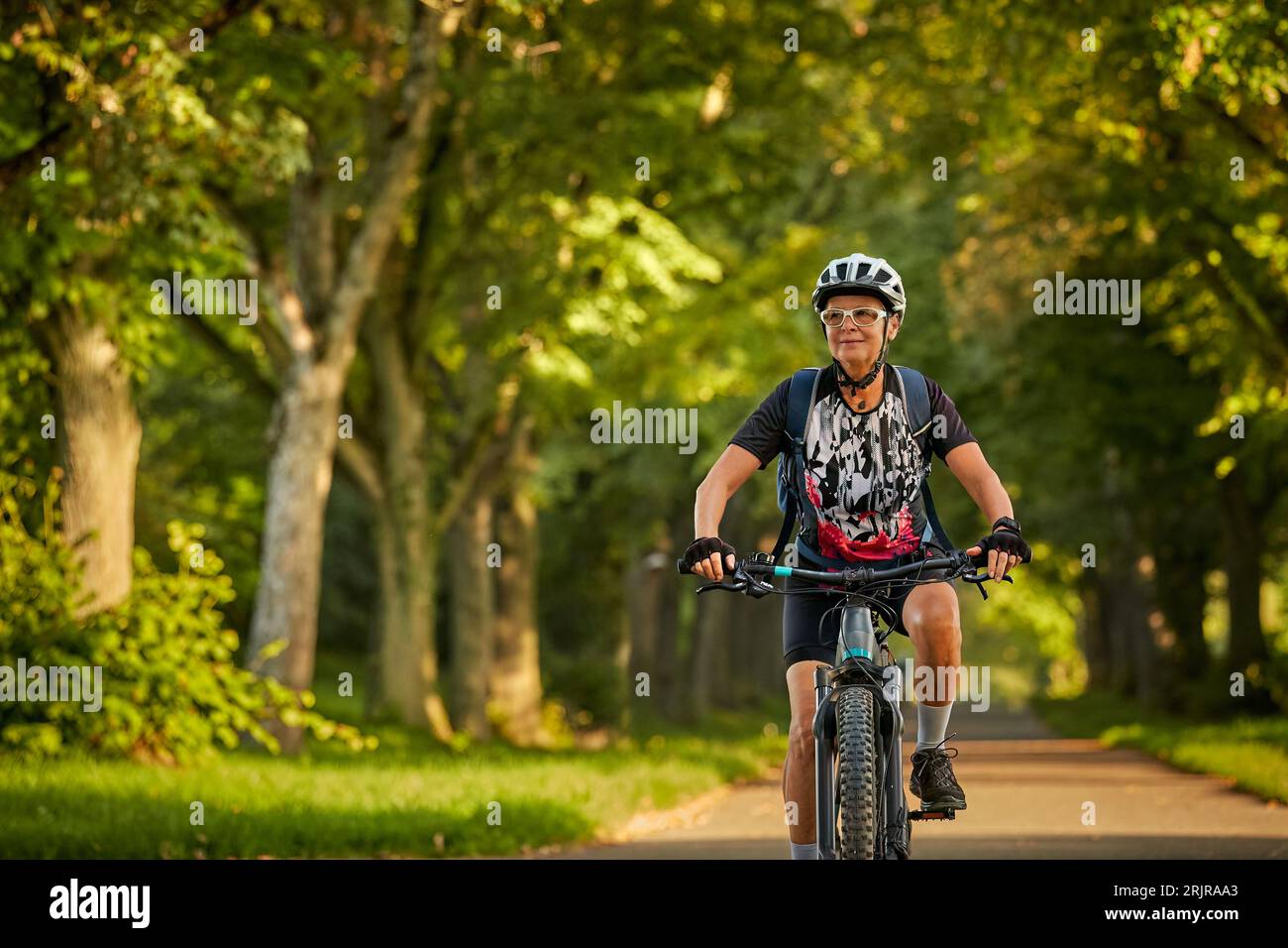 Attraktive Seniorenradfahrerin mit ihrem E-Mountainbike in einer schönen alten Eichen- und Kastanienallee in Ludwigsburg, Baden-Württemberg, GE Stockfoto