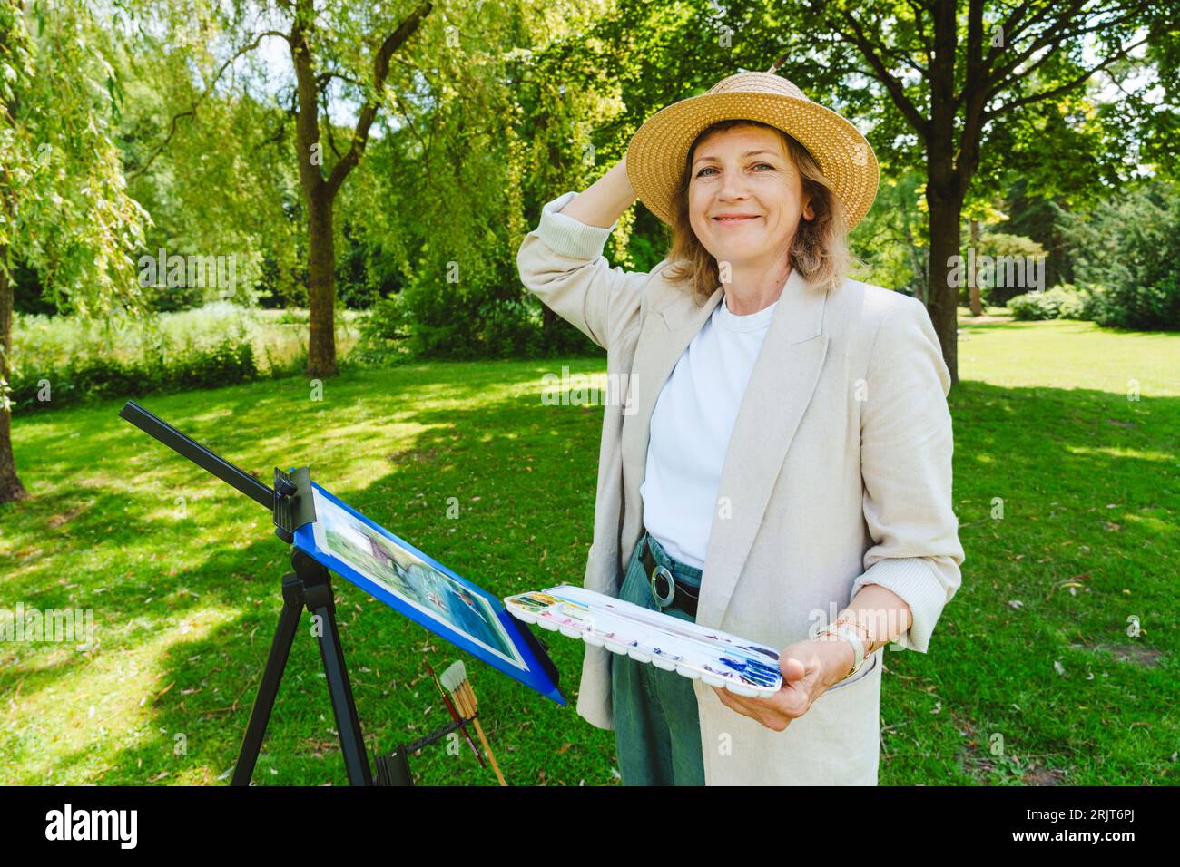 Lächelnde reife Frau, die im Park einen Hut mit Aquarellfarben trägt Stockfoto