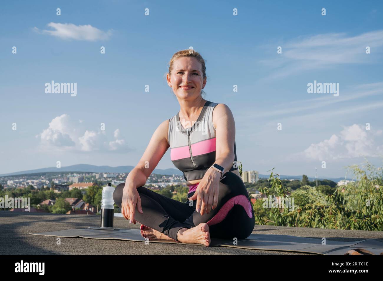 Lächelnde Frau, die auf der Yogamatte auf dem Dach sitzt Stockfoto
