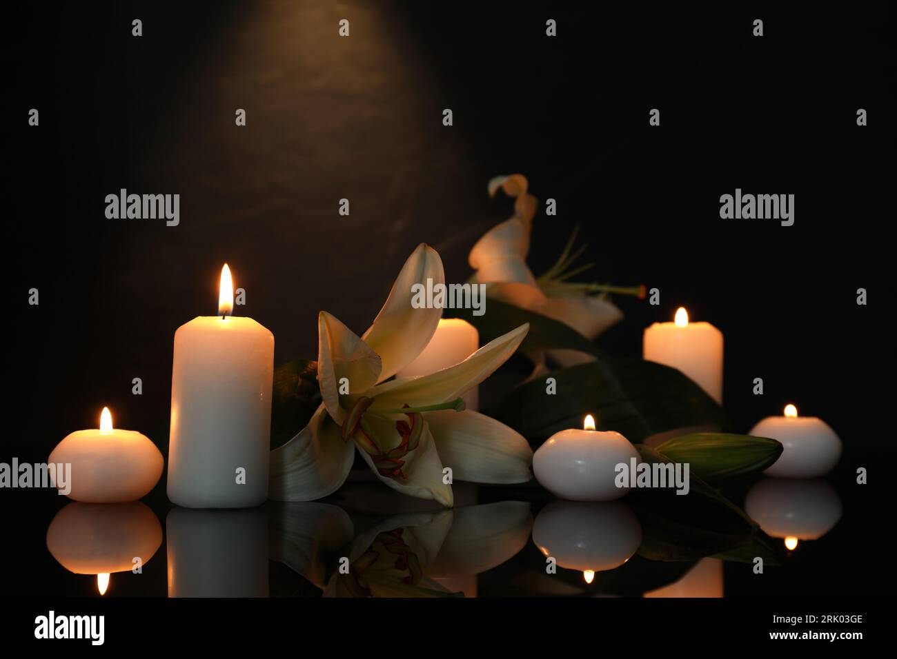 Weiße Lilien und brennende Kerzen auf schwarzer Spiegeloberfläche bei Dunkelheit, Platz für Text. Begräbnissymbole Stockfoto