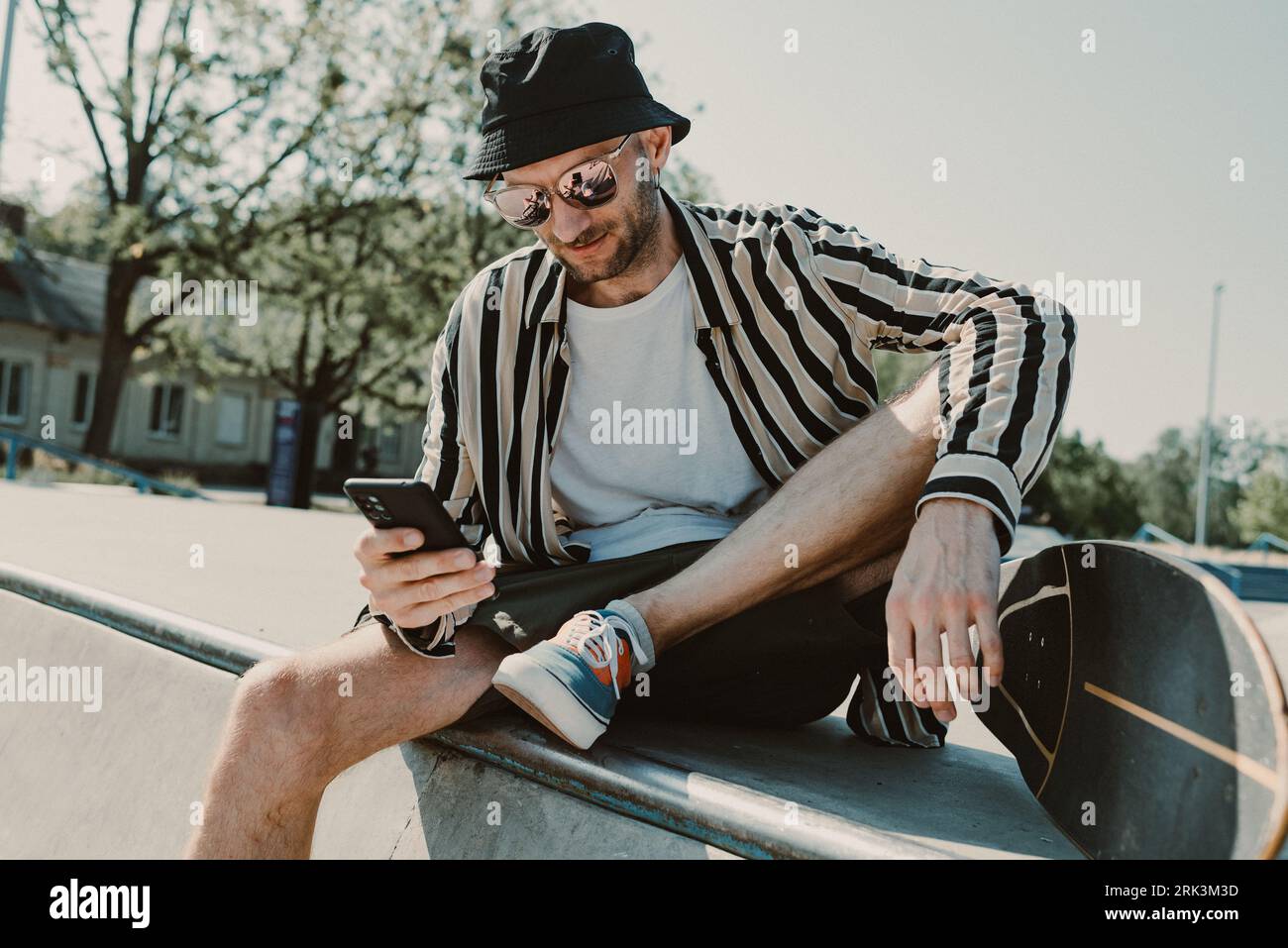 Attraktiver, eleganter Erwachsener, der an der Skatepark-Rampe mit einem Skateboard sitzt, mit dem Handy telefoniert und sich ausruht Stockfoto