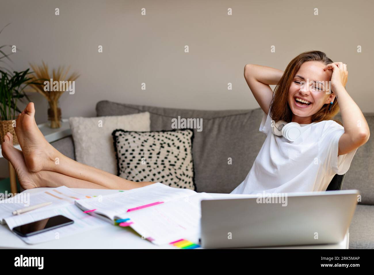 Junge lachende Studentin im weißen T-Shirt, die zu Hause lernt, E-Learning auf dem Laptop nutzt und Spaß hat. Stockfoto