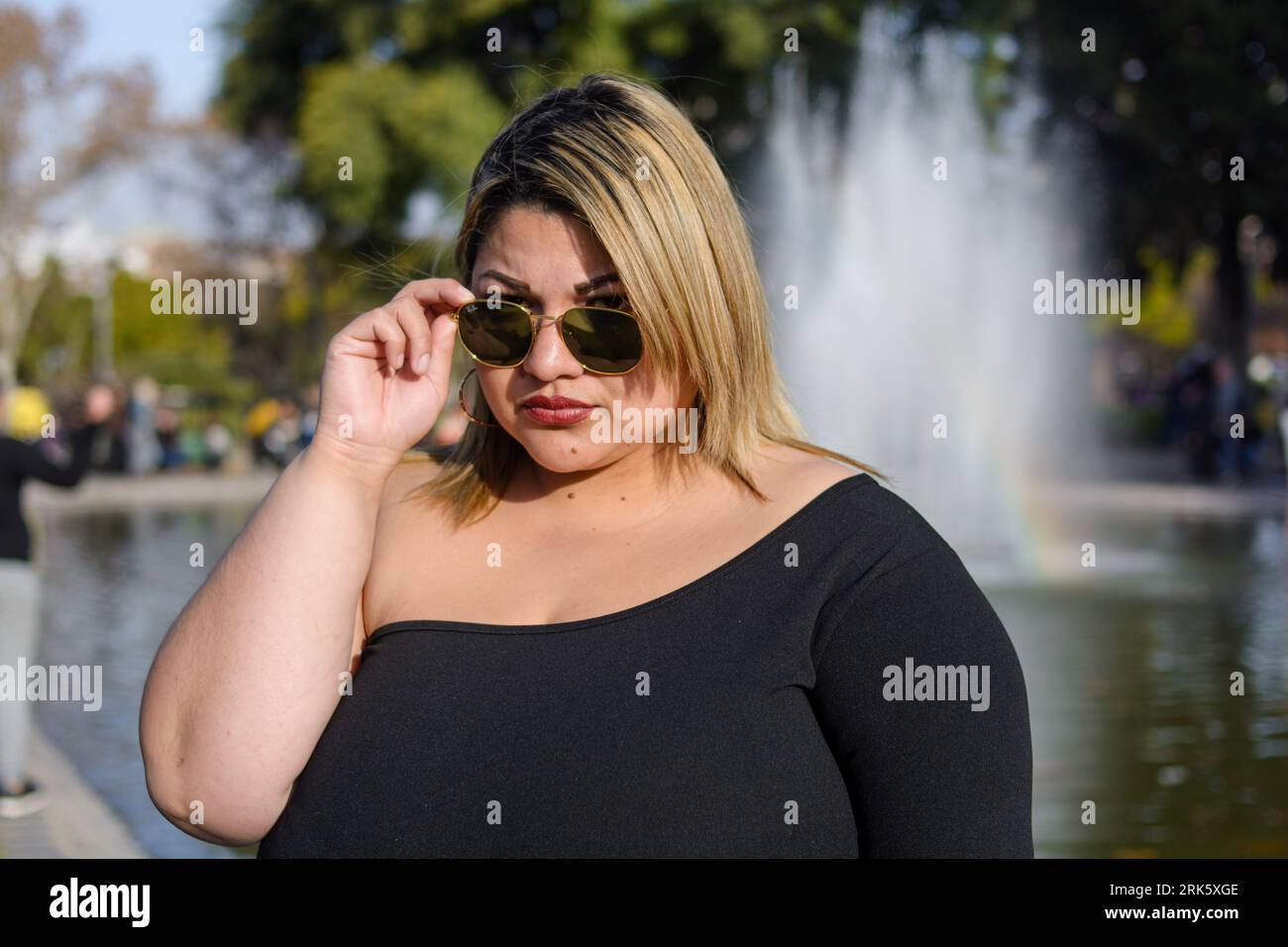 Porträt der jungen blonden Plus Größe modische Frau der argentinischen Latina Ethnicity, die draußen im öffentlichen Park steht und sich mit Sonnenbrille mit W posiert Stockfoto