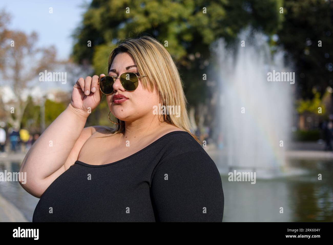 Porträt der jungen blonden Plus Größe modische Frau der argentinischen Latina Ethnicity, die draußen im öffentlichen Park steht und sich mit Sonnenbrille mit W posiert Stockfoto