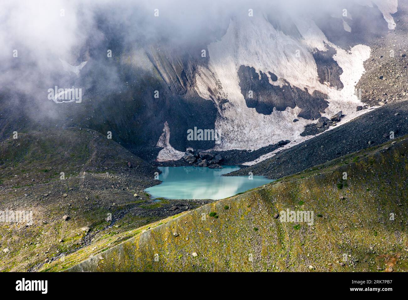 Ein milchig-turquoisblauer Bergsee (möglicherweise einer der Abudelauri-Seen) liegt unter tief hängenden Wolken am Fuße von Schneefeldern auf dem Aufstieg zum Chaukhi-Pass in der Munizipalität Duscheti, Region Mtskheta-Mtianeti, Georgien. Dunkle Felsbrocken liegen im Schnee am oberen Ende des Sees. Links reichen Geröllhänge bis zum Ufer hinab. Vorne fällt ein grüner Hang mit Steinen ab. Die drei Abudelauri-Seen (georgisch აბუდელაურის ტბები), der Grüne, der Blaue und der Weiße See, liegen zwischen dem Pass und dem Dorf Roshka. Der Pass liegt auf etwa 3.340 Metern Höhe und führt nach Juta in der Munizipalität Kazbegi. Stockfoto