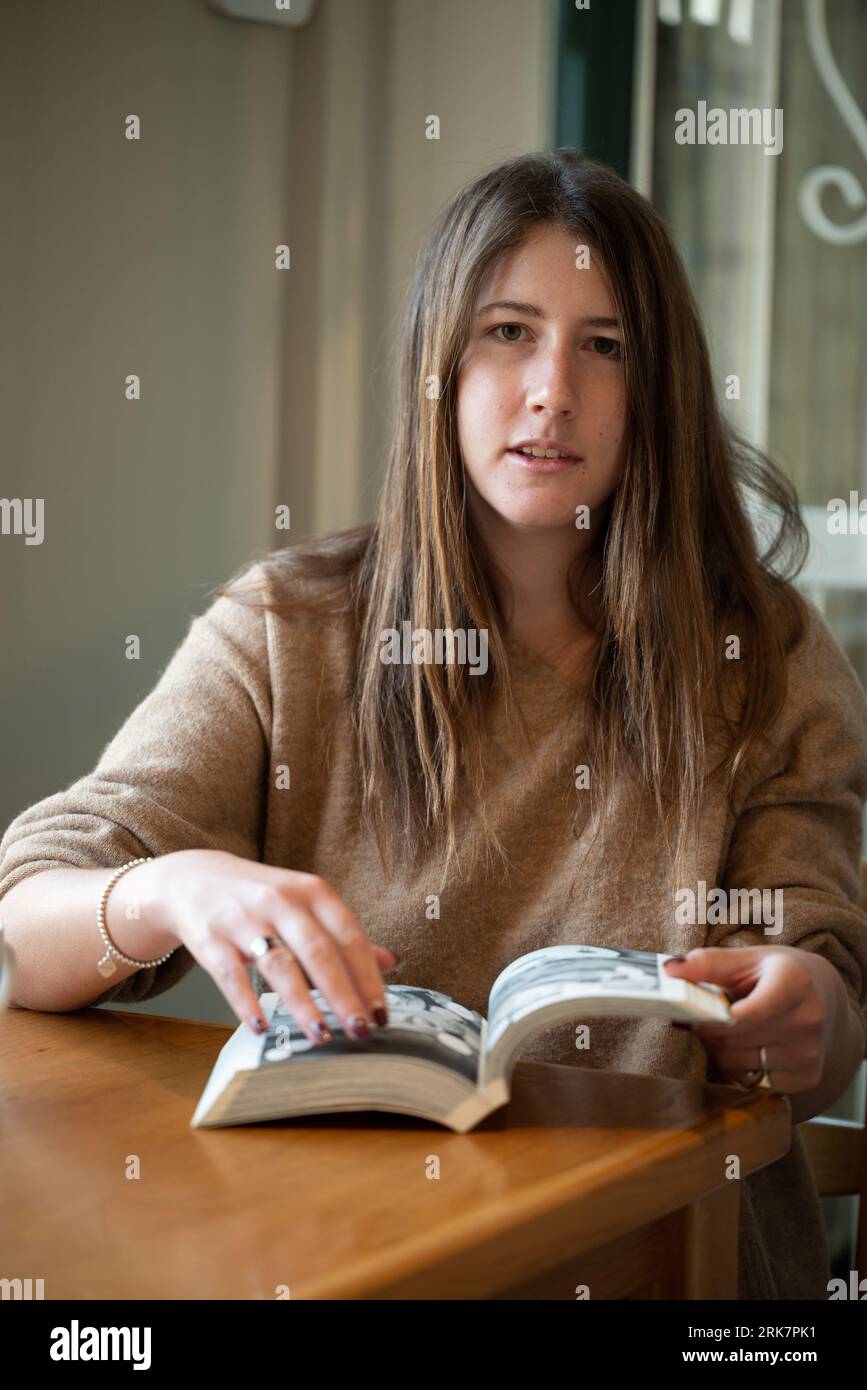 Eine fröhliche brünette Frau in einem braunen Jersey sitzt an einem Küchentisch, eingetaucht in ein Manga-Buch Stockfoto