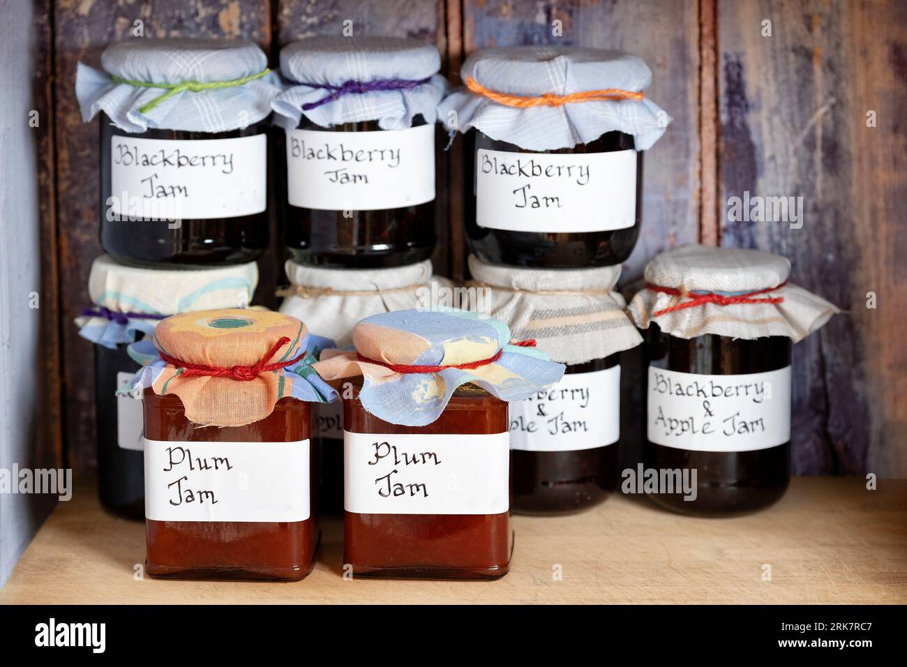 Verschiedene Gläser mit hausgemachter Pflaume, Brombeermarmelade, handgeschriebenen Etiketten und Stoffauflagen auf einem Regal in einem rustikalen Holzschrank Stockfoto
