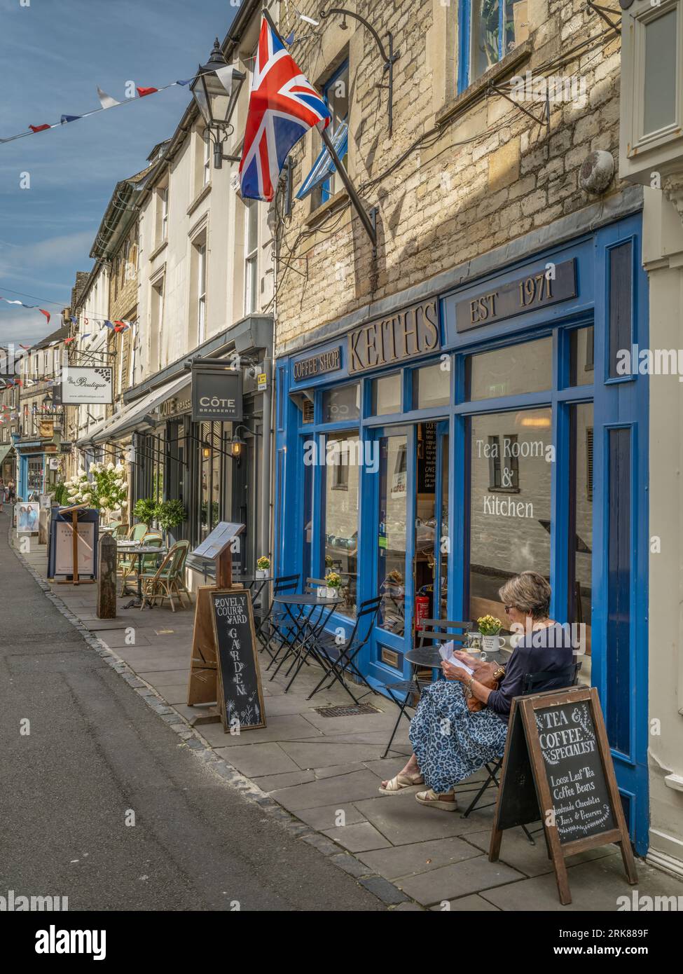 Eine Dame sitzt und genießt die Sommersonne bei einem Kaffee vor einem Straßencafé in der Black Jack Street, Cirencester, Gloucestershire. Stockfoto