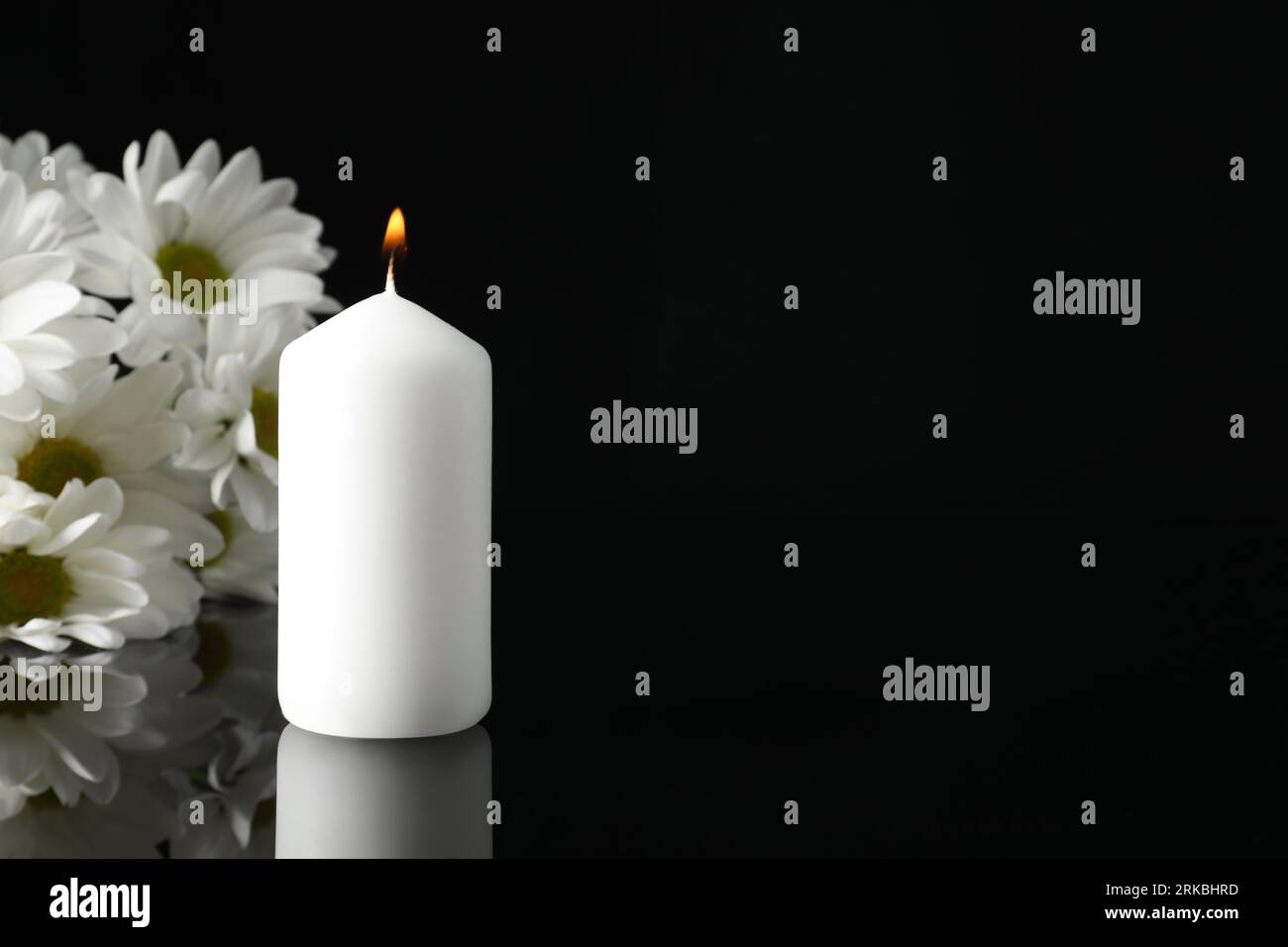 Brennende Kerze und weiße Chrysantheme-Blüten auf schwarzer Spiegeloberfläche in Dunkelheit, Raum für Text. Beerdigungssymbole Stockfoto