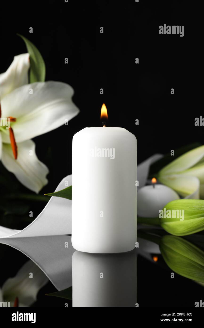 Weiße Lilien und brennende Kerze auf schwarzer Spiegeloberfläche im Dunkeln, Platz für Text. Beerdigungssymbole Stockfoto