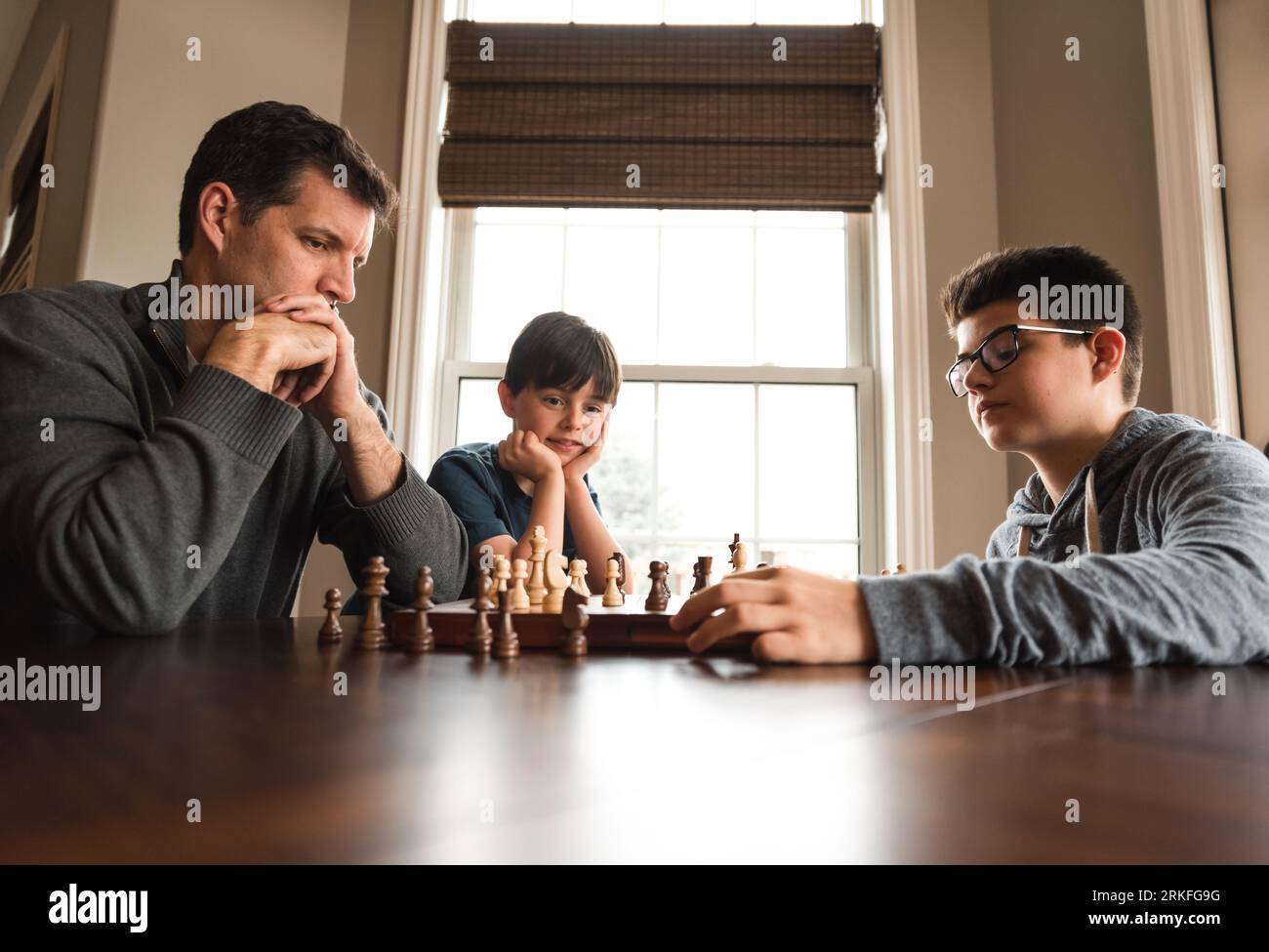 Vater und Sohn spielen Schach an einem Tisch, während der kleine Bruder zusieht. Stockfoto