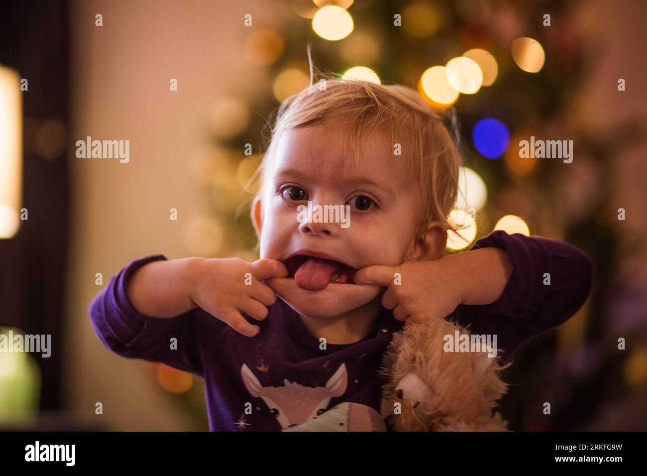 Kleinkind macht dummes Gesicht vor der Kamera mit weihnachtsbokeh im Hintergrund Stockfoto