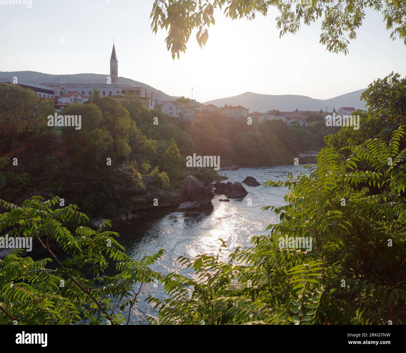 Blick zwischen üppigen Bäumen am Fluss Neretva in Mostar, mit einem Dorf und einer Moschee am Flussufer, Bosnien und Herzegowina, 22. August 2023. Stockfoto